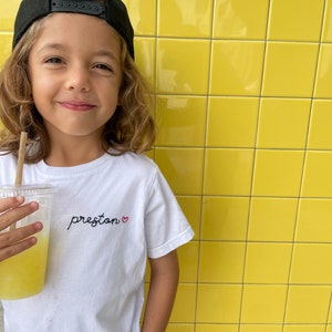 May include: A young person wearing a black baseball cap and a white t-shirt with the word "preston" embroidered on it in black with a small red heart. The person is holding a clear plastic cup with a straw and a yellow beverage.