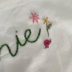 May include: Embroidered word "nie" in green thread with pink and yellow flowers and a pink rose.
