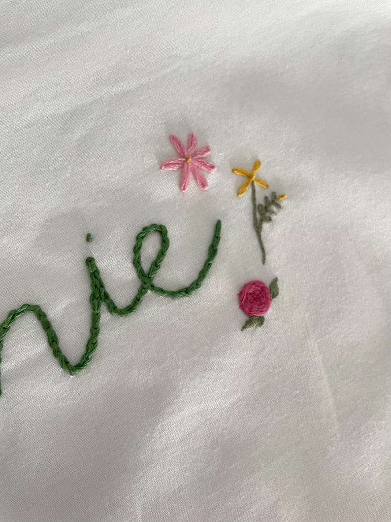 May include: Embroidered word "nie" in green thread with pink and yellow flowers and a pink rose.