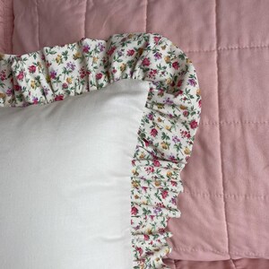 May include: A white pillow with a ruffled edge. The ruffle is made of a white fabric with a small floral print in shades of pink, yellow, and green. The pillow is sitting on a pink quilted fabric.