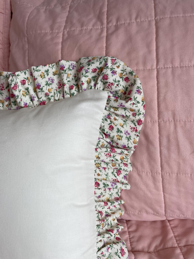 May include: A white pillow with a ruffled edge. The ruffle is made of a white fabric with a small floral print in shades of pink, yellow, and green. The pillow is sitting on a pink quilted fabric.