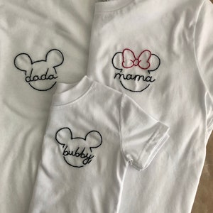May include: Three white t-shirts with embroidered Mickey Mouse ears and the words "dada", "mama", and "bubby".