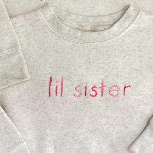 May include: A light gray sweatshirt with the embroidered text "lil sister" in shades of pink. The sweatshirt has a crew neck and long sleeves with ribbed cuffs. The fabric has a speckled texture.