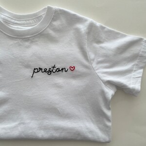 May include: White t-shirt with black embroidered text that says "preston" followed by a small red heart.