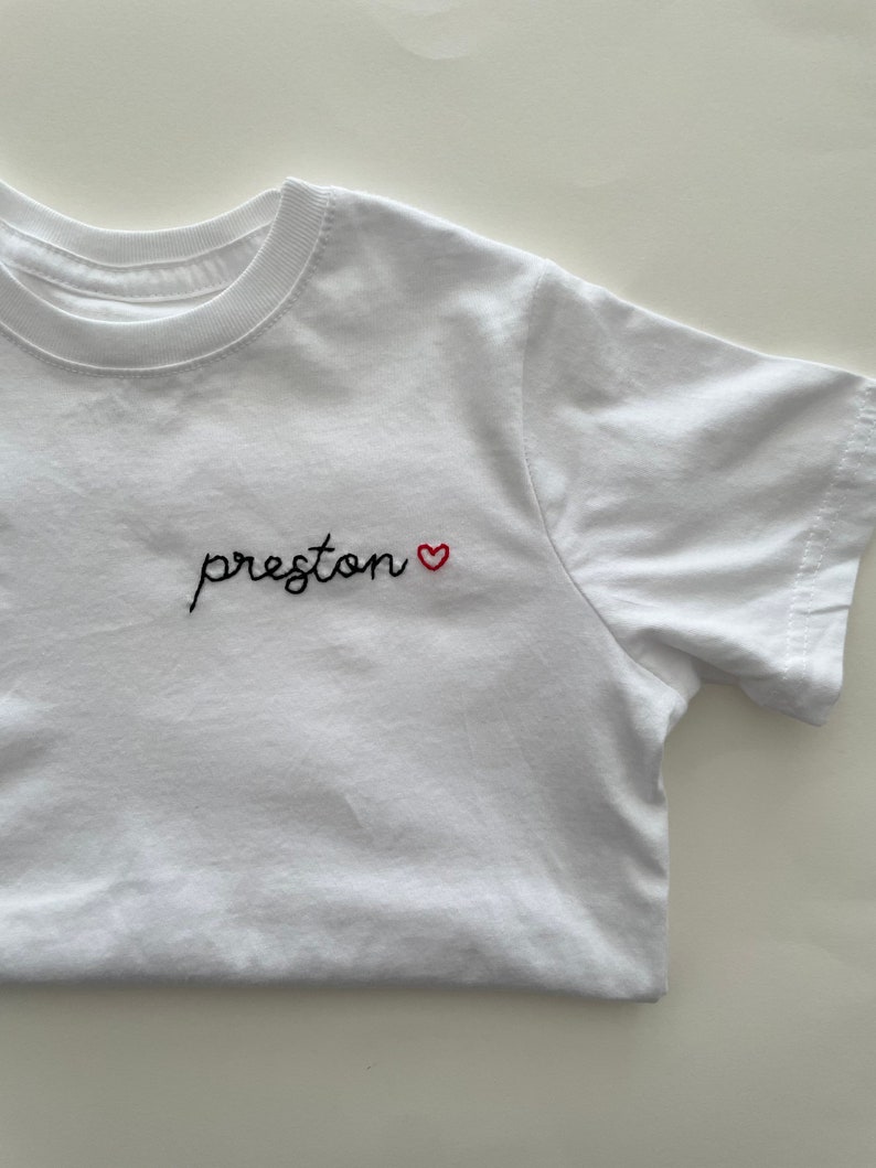 May include: White t-shirt with black embroidered text that says "preston" followed by a small red heart.
