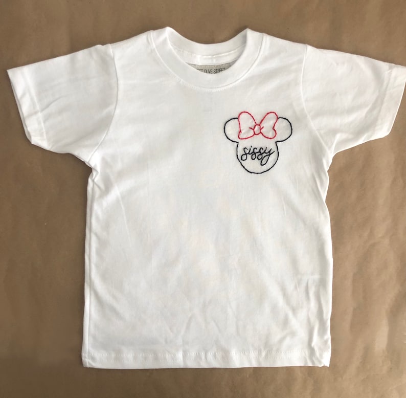 May include: White t-shirt with a black and red embroidered Minnie Mouse design. The design includes a bow and the word "sissy".