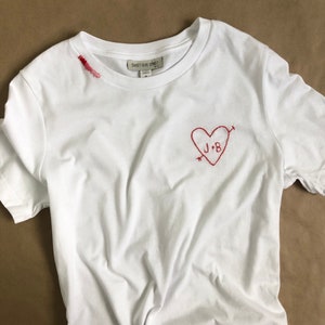 Puede incluir: Camiseta blanca con un corazón bordado en rojo y las iniciales "J+B" en su interior. La camiseta tiene una pequeña mancha roja en el cuello.