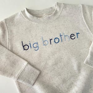 Op de afbeelding: Een lichtgrijs sweatshirt met de woorden "grote broer" geborduurd in blauwe draad.