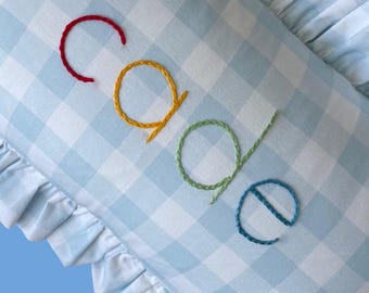 May include: A light blue and white gingham pillow with the name "CAOE" embroidered in red, yellow, green, and blue thread. The pillow has a ruffled edge.