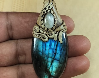 Puede incluir: Un colgante artesanal con una piedra de labradorita ovalada pulida con iridiscencia azul y verde. La piedra está rematada con un elemento tallado de forma orgánica, de color marrón claro, y una pequeña piedra ovalada blanca. El colgante se sostiene en una mano.