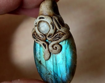 Puede incluir: Colgante artesanal con una piedra de labradorita ovalada pulida con tonos iridiscentes azules y verdes. La piedra está rematada con un diseño de madera tallada, que muestra detalles intrincados y un pequeño acento de piedra blanca redonda.