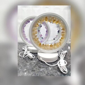 May include: Two white circular lamps with a ring of yellow and purple gemstones around the perimeter. The lamps are on a white base and have a cord.