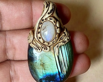 Puede incluir: Un colgante artesanal con una gran piedra ovalada de labradorita con iridiscencia azul y verde. El colgante está rematado con un elemento decorativo tallado, de color marrón claro, y una piedra lunar ovalada más pequeña. El colgante se sostiene en una mano.