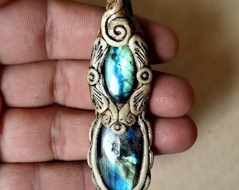 Puede incluir: Un colgante artesanal con dos piedras ovaladas de labradorita con tonos iridiscentes azules y verdes. Las piedras están engastadas en un marco beige, tallado y ornamentado con espirales y detalles en forma de hojas, creando una pieza artística única.
