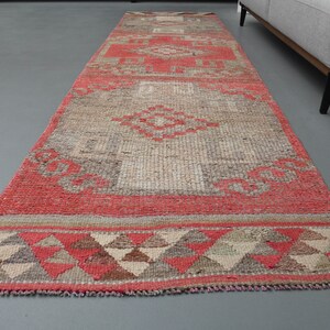 Könnte beinhalten: Ein langer, rechteckiger Teppich mit einem geometrischen Muster in Rot, Beige und Braun. Der Teppich hat einen Fransenrand und ist aus einem gewebten Material gefertigt.