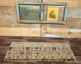 May include: A vintage rug with a beige background and colorful geometric patterns. The rug is laid out on a wooden floor in front of a wall with two framed pictures.