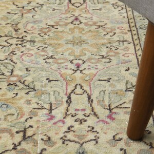 May include: A close-up of a beige rug with a floral pattern. The rug has a brown wooden leg resting on it.
