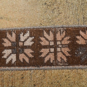 Puede incluir: Primer plano de una sección de una alfombra con un borde marrón con un patrón floral repetido en beige y marrón claro. El fondo es beige texturizado.