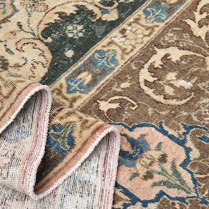 May include: Close-up of a vintage rug with a detailed floral pattern. The rug features a colour palette of beige, brown, blue, and pink. The edges are folded, revealing the rug's backing. The design includes floral and geometric elements.