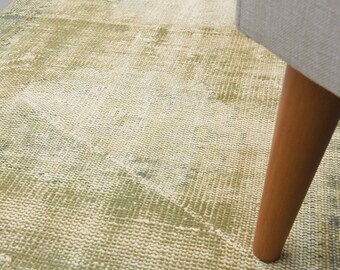 May include: A close-up of a distressed, vintage-style rug in faded green and cream tones. The rug's texture is visible, with a wooden furniture leg in the foreground, suggesting a living room or home decor setting.