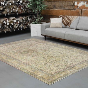 May include: A vintage cream-colored rug with a floral pattern and a faded pink border. The rug is laid out on a gray floor in a room with a gray sofa and a wooden wall.