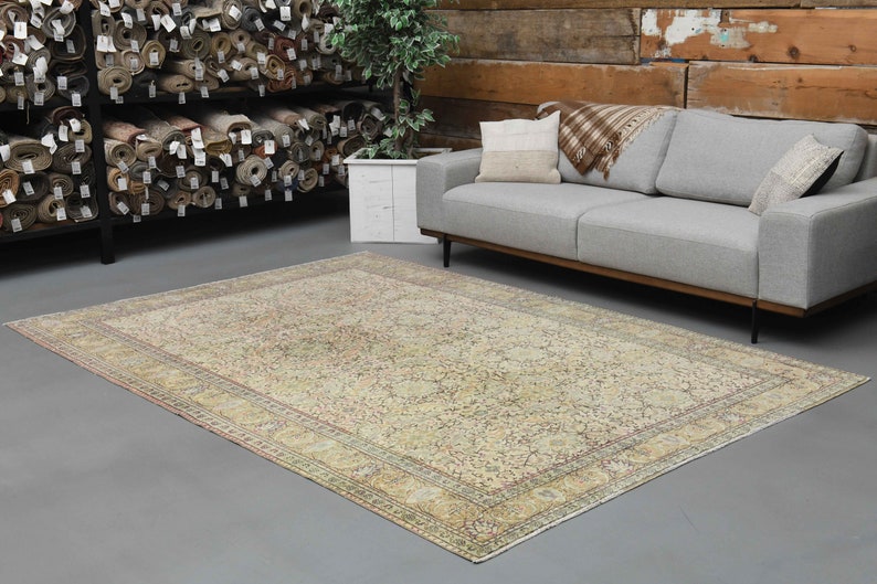 May include: A vintage cream-colored rug with a floral pattern and a faded pink border. The rug is laid out on a gray floor in a room with a gray sofa and a wooden wall.