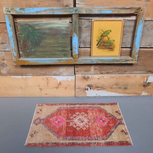 May include: A small, rectangular rug with a red, pink, and beige pattern. The rug has a distressed look and is made of woven fibers. The rug is sitting on a gray floor in front of a wooden wall with two framed pictures.
