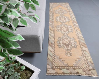 May include: A vintage runner rug with a light peach background and intricate geometric patterns in shades of brown, gray, and green. The rug has a fringed edge and is perfect for adding a touch of bohemian style to any room.