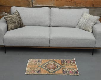 Könnte beinhalten: Ein hellgraues Sofa mit zwei Kissen, zwei dekorativen Kissen und einem kleinen, gemusterten Teppich. Der Teppich hat ein geometrisches Design mit orangefarbenen, braunen und rosa Akzenten. Das Sofa hat eine dunkle Holzbasis und schwarze Beine.