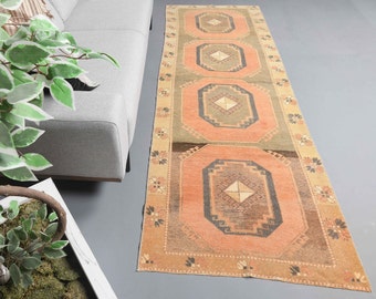 May include: A long, rectangular rug with a geometric pattern in shades of brown, green, and pink. The rug has a fringe border and is made of wool.