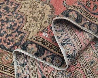 May include: Vintage-style rug with a distressed appearance. The rug features a combination of pink, beige, and navy blue colors. The design includes floral and geometric patterns, with a rolled-up edge revealing the rug's layered construction. The rug is suitable for home decor.