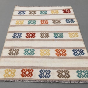 May include: A white rug with a geometric pattern in shades of blue, green, yellow, orange, and brown. The rug has a fringed border.