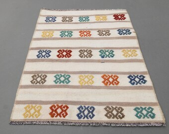 May include: A white rug with a geometric pattern in shades of blue, green, yellow, orange, and brown. The rug has a fringe border.
