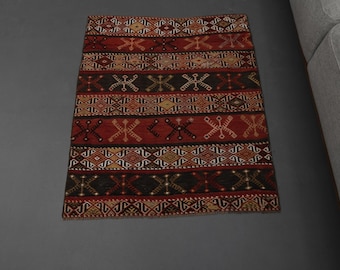 May include: A rectangular, patterned rug with a dark red and brown color scheme. The rug features geometric designs in cream, gold, and black, arranged in horizontal stripes. The rug is placed on a gray surface.