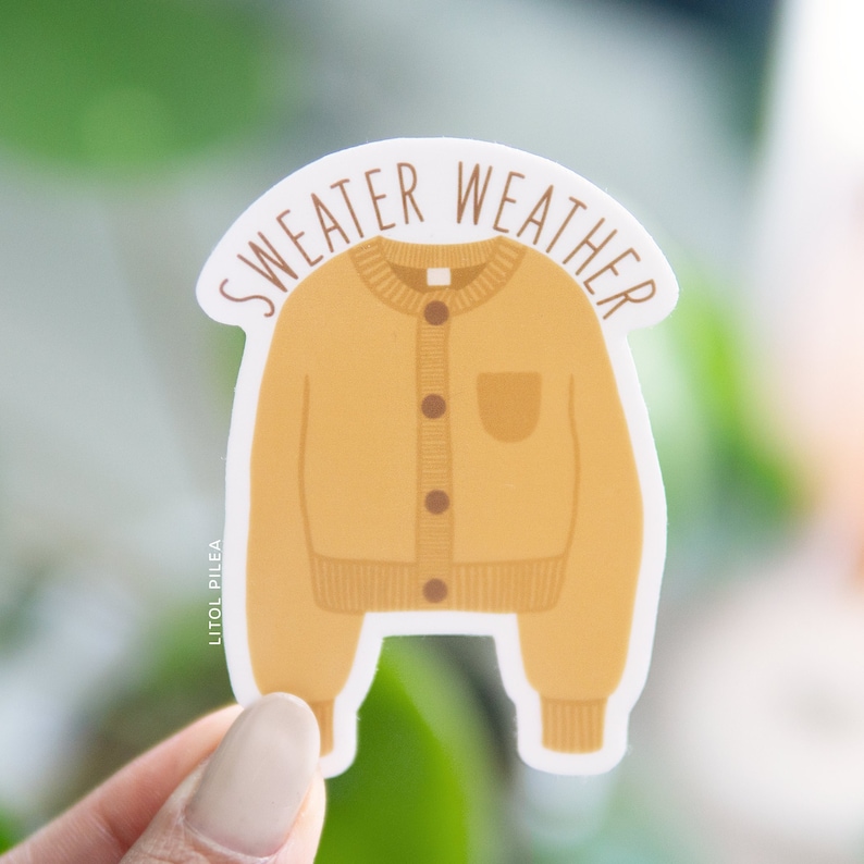 Pu&ograve; includere: Un adesivo con un cardigan giallo con il testo "sweater weather" in lettere marroni. L'adesivo &egrave; su uno sfondo bianco.