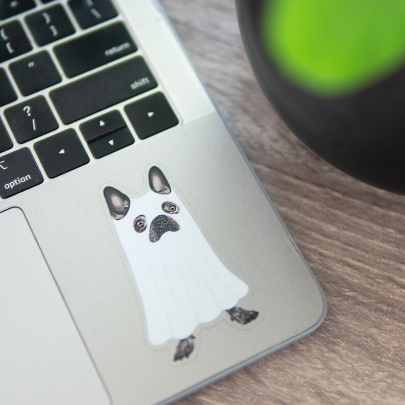 May include: A white ghost sticker with a black and white French bulldog face peeking out from the bottom. The sticker is on a silver laptop.