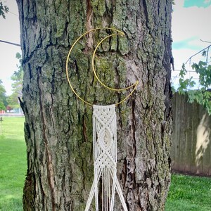 May include: A handmade macram&eacute; plant hanger with a terracotta pot suspended from a gold crescent moon-shaped metal frame. The plant holder is made of off-white rope and is attached to a tree trunk.