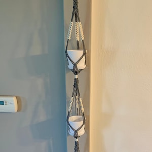May include: A macrame plant hanger with three white ceramic pots. The hanger is made of gray and cream-colored rope, with a tassel at the bottom. The plant hanger is suspended from a white hook on a wall.