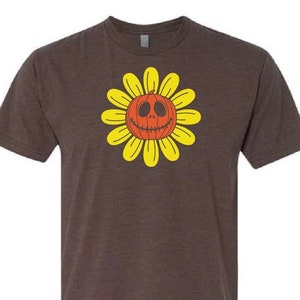 Op de afbeelding: Een bruin T-shirt met een geel zonnebloemontwerp. Het midden van de zonnebloem is een rode pompoen met een gesneden jack-o'-lantern gezicht.
