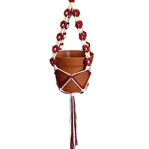 Peut inclure: Un porte-plante suspendu en macram&eacute; blanc et bordeaux avec un anneau en bois en haut. Le porte-plante a un pompon bordeaux et tient un pot en terre cuite.