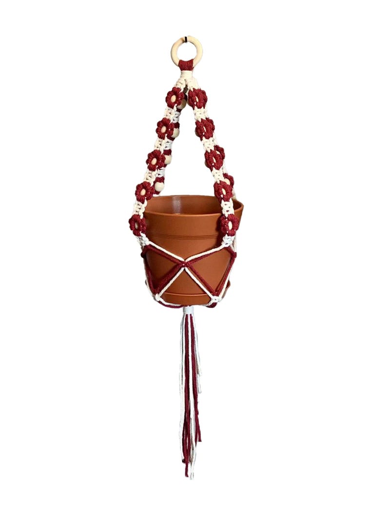 Peut inclure: Un porte-plante suspendu en macram&eacute; blanc et bordeaux avec un anneau en bois en haut. Le porte-plante a un pompon bordeaux et tient un pot en terre cuite.