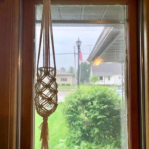 May include: A white macrame plant hanger with a glass jar hanging from it. The hanger is hanging in a window with a view of a green lawn and trees.
