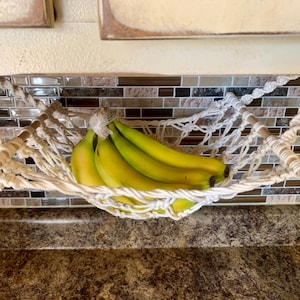 Może przedstawiać: Biały hamak na banany z makramy, trzymający pęczek zielono-ż&oacute;łtych banan&oacute;w. Hamak zawieszony jest między dwoma drewnianymi kołkami. Tło przedstawia kafelkową osłonę i blat.