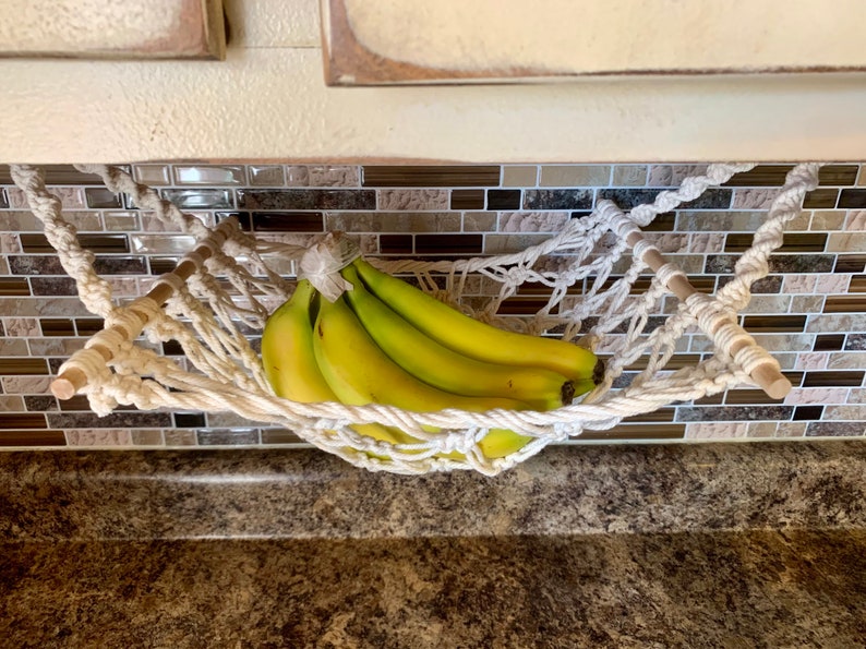 Może przedstawiać: Biały hamak na banany z makramy, trzymający pęczek zielono-ż&oacute;łtych banan&oacute;w. Hamak zawieszony jest między dwoma drewnianymi kołkami. Tło przedstawia kafelkową osłonę i blat.