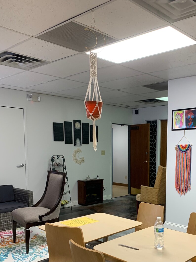 May include: A macrame plant holder with a terracotta pot suspended from a gold crescent moon-shaped fixture. The plant holder is made of off-white woven rope. The room has a modern design with neutral colours and a colourful rug.