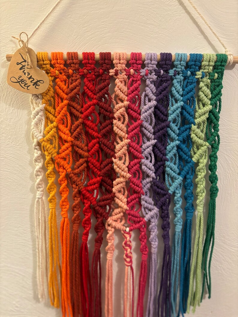 Op de afbeelding: Een regenboogkleurige macram&eacute; wandhanger met een houten lat aan de bovenkant en een hartvormig label met de tekst 'Bedankt'.