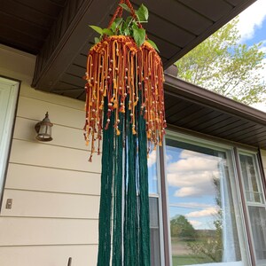 K&ouml;nnte beinhalten: Ein Makramee-Pflanzenh&auml;nger in Gr&uuml;n, Orange und Gelb. Der H&auml;nger ist mit kleinen wei&szlig;en Perlen verziert und hat eine lange Franse. Eine Topfpflanze h&auml;ngt im Makramee.