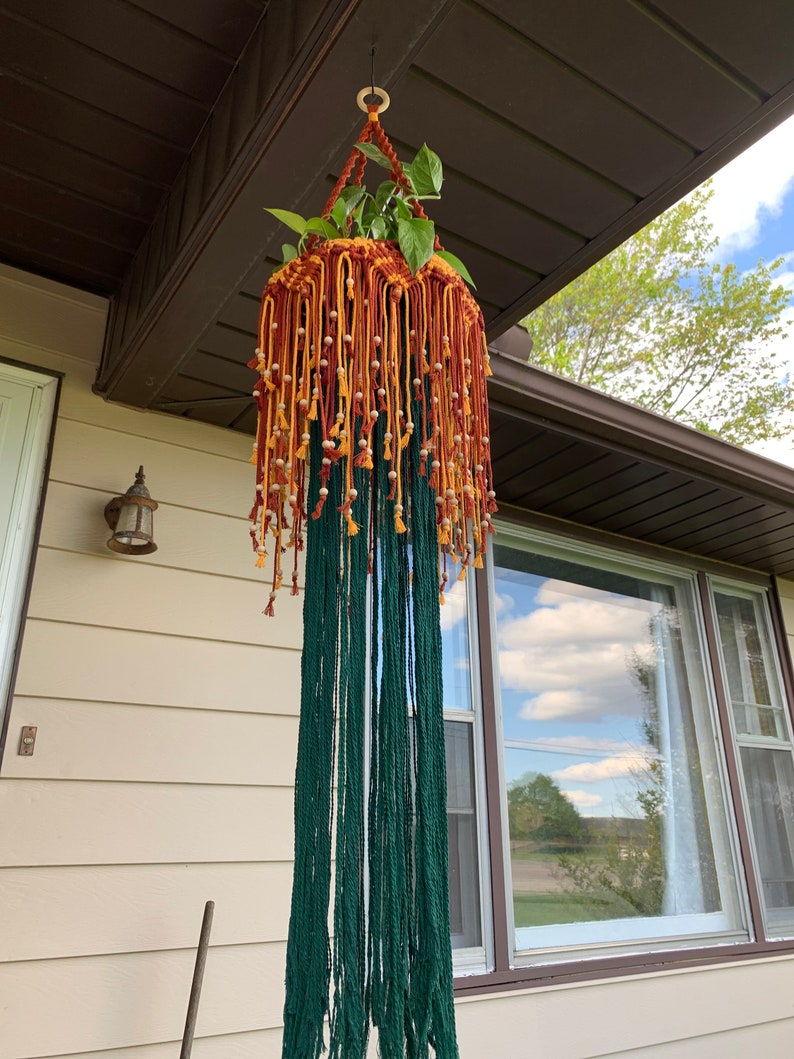 K&ouml;nnte beinhalten: Ein Makramee-Pflanzenh&auml;nger in Gr&uuml;n, Orange und Gelb. Der H&auml;nger ist mit kleinen wei&szlig;en Perlen verziert und hat eine lange Franse. Eine Topfpflanze h&auml;ngt im Makramee.