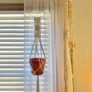 May include: A macram&eacute; plant hanger with a terracotta pot suspended from a gold crescent moon-shaped ring. The hanger is made of off-white rope and hangs in front of a sheer white curtain. Additional macram&eacute; decor is visible.