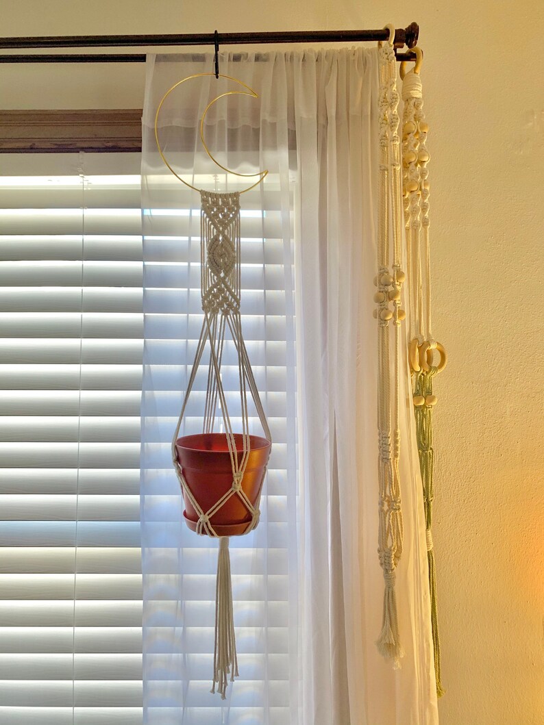 May include: A macram&eacute; plant hanger with a terracotta pot suspended from a gold crescent moon-shaped ring. The hanger is made of off-white rope and hangs in front of a sheer white curtain. Additional macram&eacute; decor is visible.